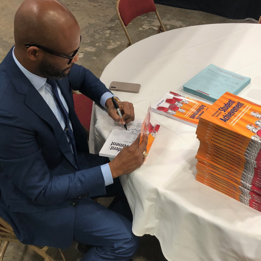 Dr. Dickey sitting on a table signing an autograph on books about student achievement